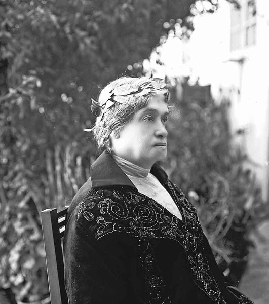 Fotografía en blanco y negro de Adela Zamudio anciana, sentada de perfil en un jardín, con corona de laurel sobre el cabello cano y abrigo oscuro bordado.