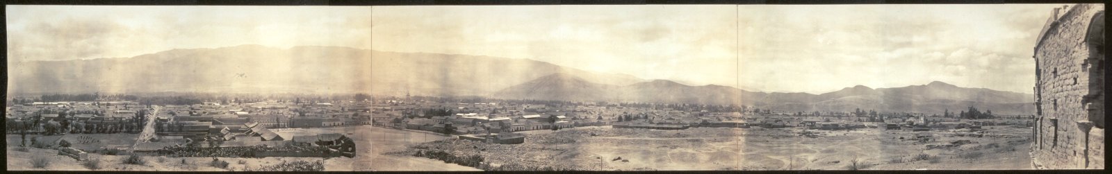 Panorámica en sepia de Cochabamba en 1915: casas bajas con tejados y algunos árboles en un valle llano, con una larga cadena de montañas al fondo y un muro de adobe en ruinas en el extremo derecho.