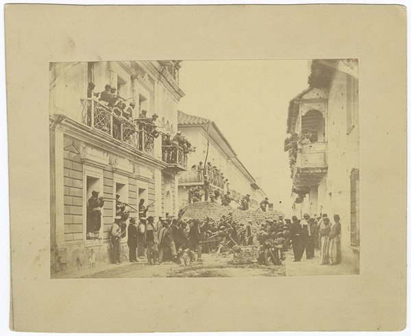 Fotografía antigua desvaída montada en cartón: calle con casas de balcones, llena de gente agolpada; espectadores asomados a los balcones y ventanas.