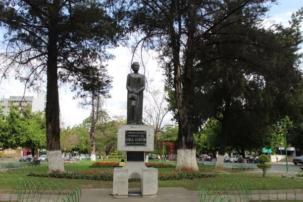 Estatua oscura de Adela Zamudio de pie sobre un pedestal blanco con una placa, en un paseo arbolado con césped y flores, bajo un cielo azul.